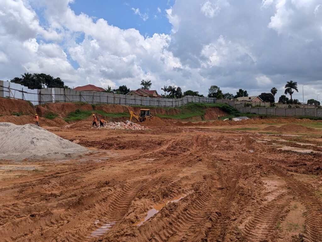 A construction site with a large, cleared dirt area under a partly cloudy sky. Several workers in orange vests are scattered