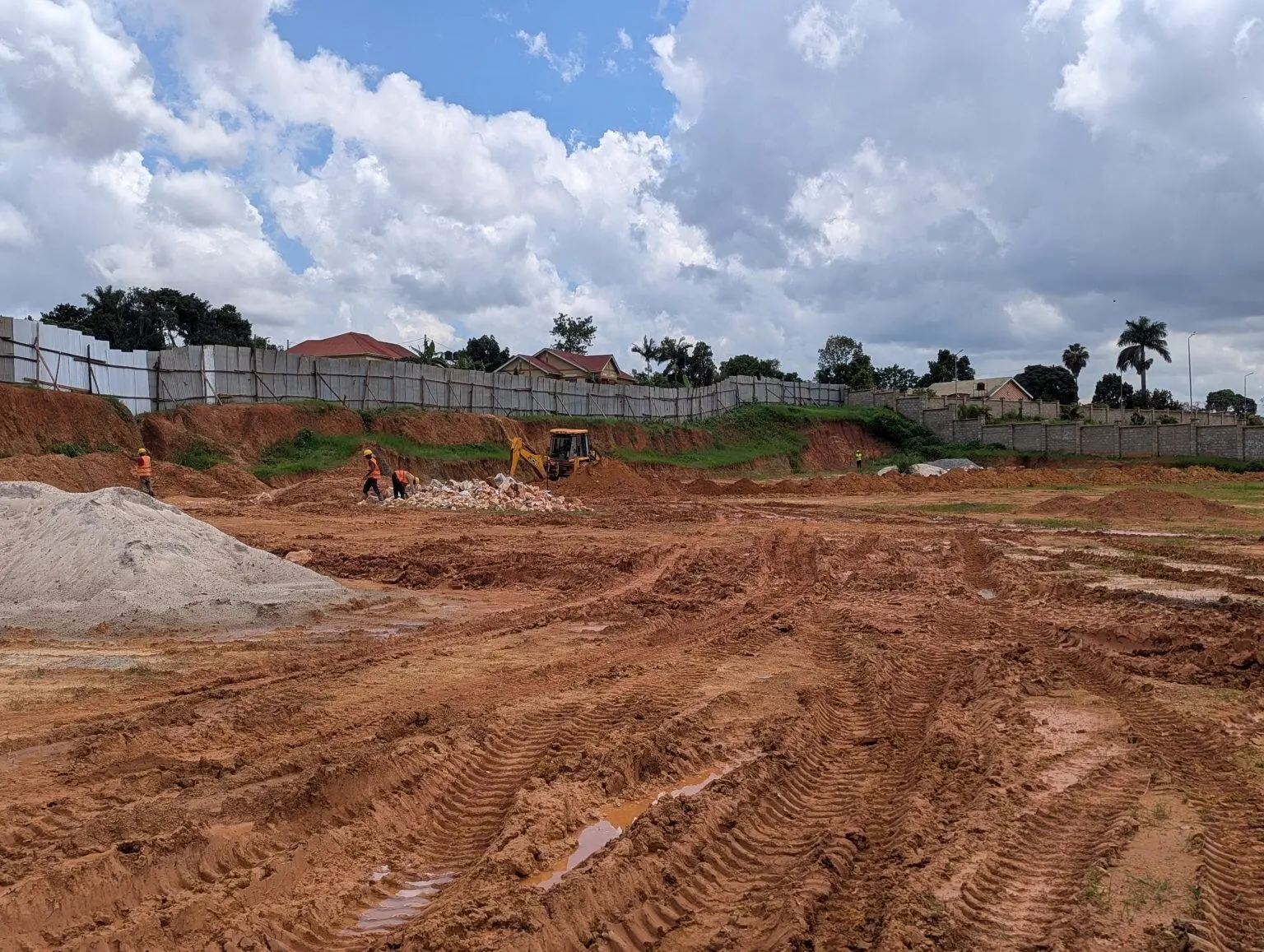 A construction site with a large, cleared dirt area under a partly cloudy sky. Several workers in orange vests are scattered