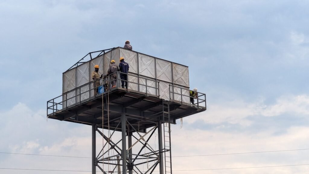 Four men wearing yellow helmets on their heads are fixing a high pressure metallic water tank.