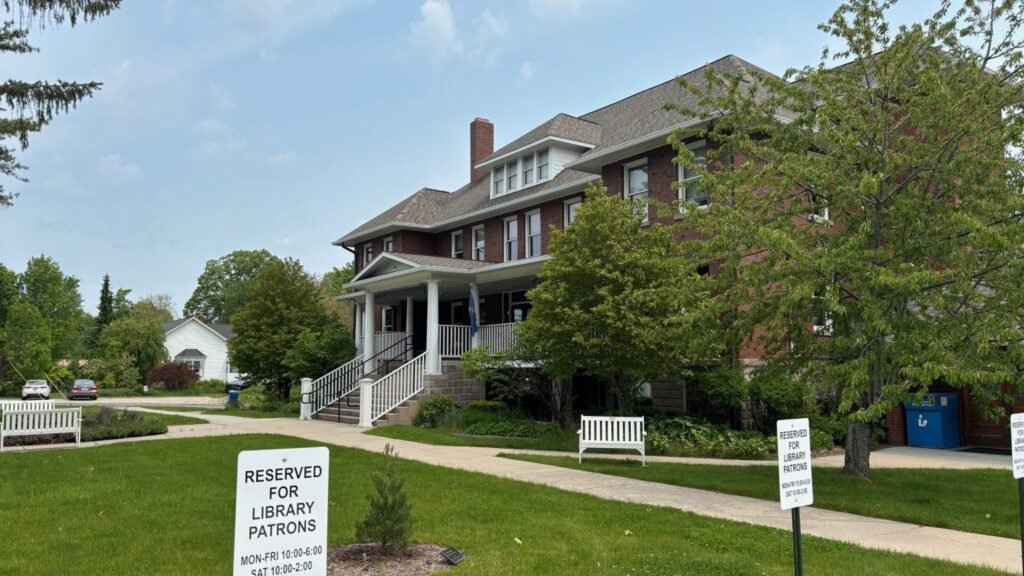 A stately old brock building with a large porch, trees and a lawn un front. In the foreground are some signs that say "Librar